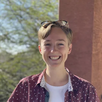 A person smiling outside with short hair and a maroon button down shirt