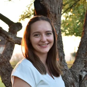 A woman in a white shirt smiling in front of trees and a sunset