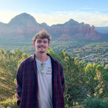 A man wearing a t-shirt and blue jacket standing in a lush valley surrounded by mountains