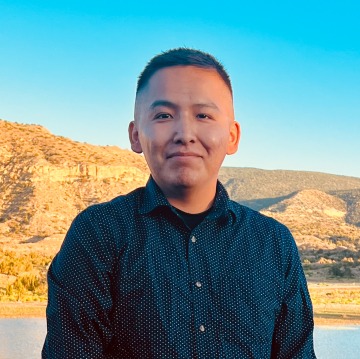 A man with cropped hair wearing a blue button-down shirt standing in front of a desert canyon