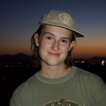 A person smiling with short hair wearing an olive baseball cap at dusk