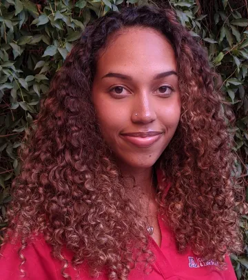 A woman with long curly brown hair wearing a red shirt, standing in front of a bush