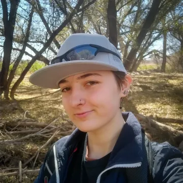 A person wearing a baseball cap with sunglasses perched on the cap brim, surrounded by trees