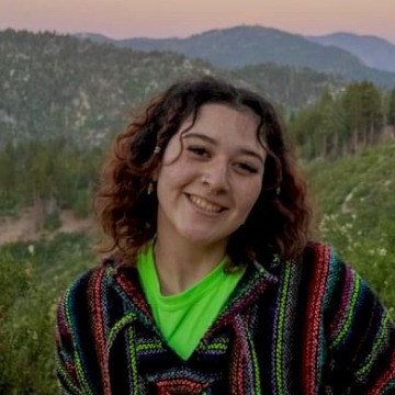 A woman smiling wearing a neon green shirt and patterned jacket, in front of a mountain sunset