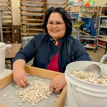 A woman with short hair and glasses wearing a denim jacket, sitting at a table with a bucket of bean seeds and a cleaning screen.