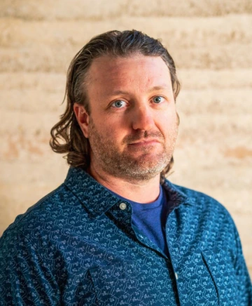A man with long hair and a short beard wearing a blue collared shirt, standing in front of an adobe wall.