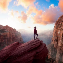 A person stands on a rocky outcrop amid mountains under a colorful sky.