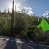 A man in a hat and green jacket pushes a walker uphill in the afternoon sun.