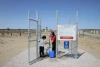 A woman and child collect water from a faucet into buckets. The faucet is protected by chain link fence in a desert.